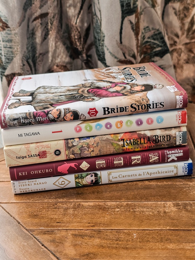 A stack of five books with various titles and colorful covers, placed on a wooden surface, against a backdrop of patterned fabric. Mangas historiques les plus passionnants à lire.