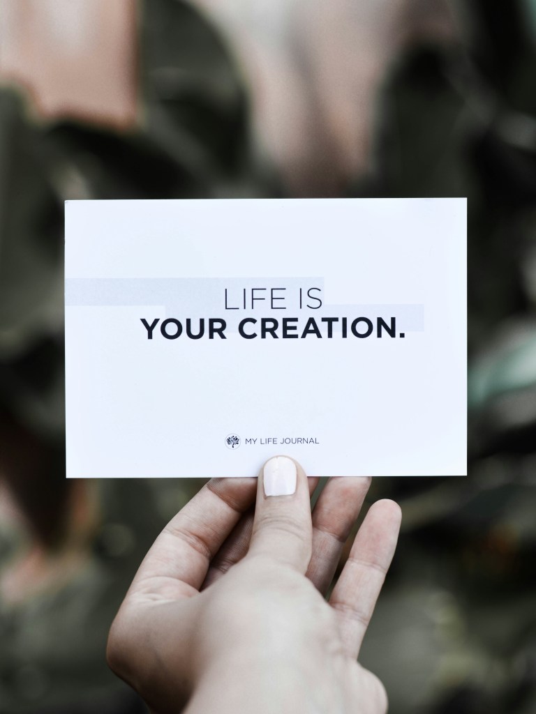 A person holding a postcard with the text 'LIFE IS YOUR CREATION.' in a stylish font, against a blurred background of green foliage.