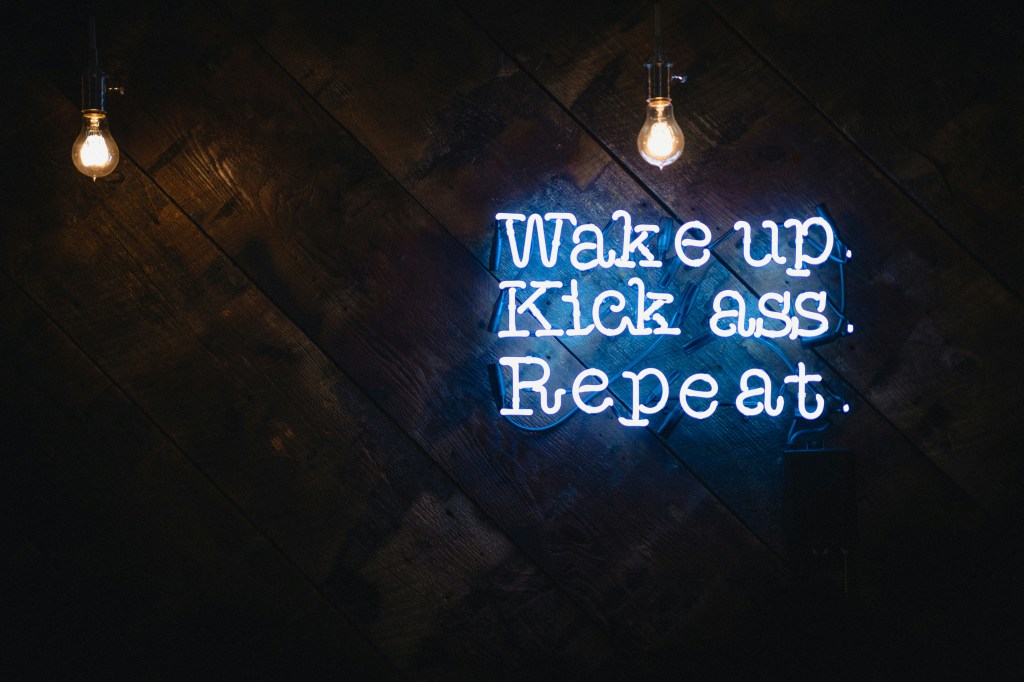 Un néon affichant le message 'Wake up. Kick ass. Repeat.' suspendu devant un fond en bois sombre.